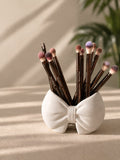 White ceramic bow-shaped brush holder with makeup brushes on a beige surface