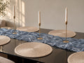Dining table styled with a baby-blue textured runner, round neutral placemats, and gold-toned candleholders.